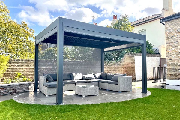 4x3m dark grey metal pergola on oval paved area set into lawn, garden brick wall behind.