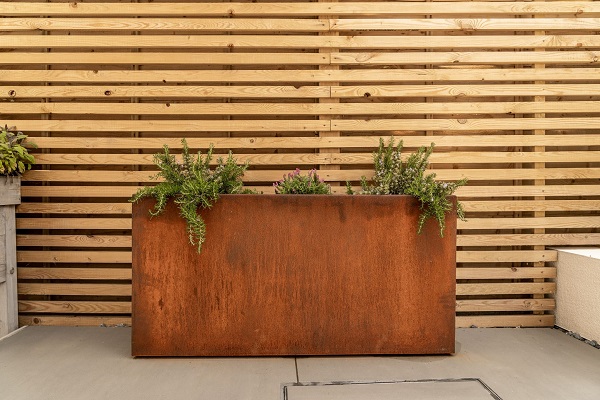 Metal trough planter in Corten Steel on paving, against open slatted fence panel. 