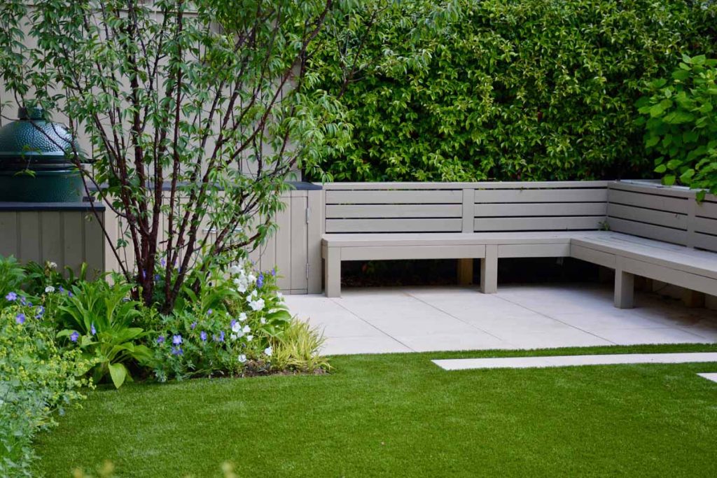 Small patio of Slab Khaki porcelain corner of hedged garden with bench on 2 sides and lawn in front.