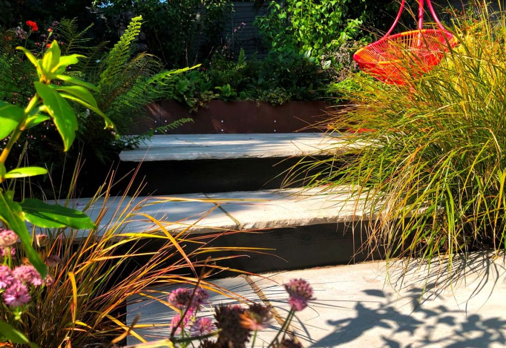 2 tumbled sandstone steps in Kandla Grey rise between grasses and ferns into shade, by VaRa Garden Design