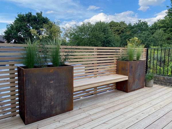 Two Corten steel trough planters provide sturdy support for a bench fixed between them against slatted fence on roof terrace.