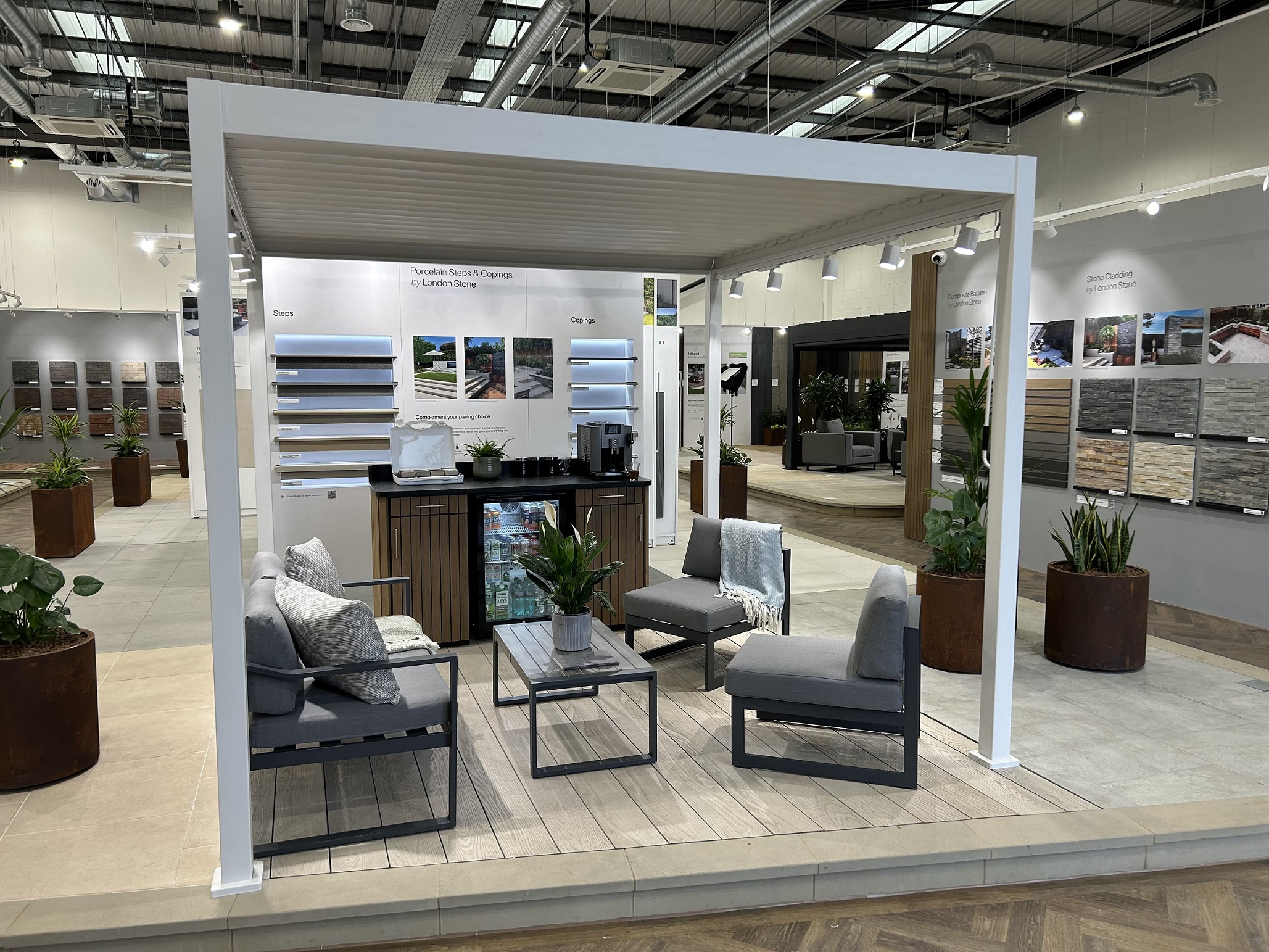 Indoor display with white pergola, furniture, coffee machine and wall displays of hard landscaping materials.