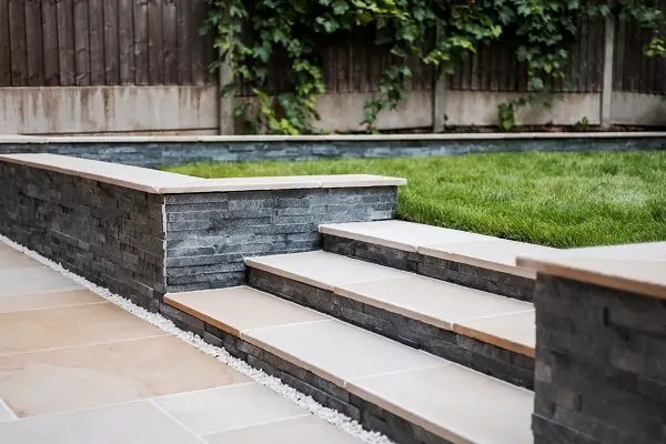 Antique cream sandstone steps from gravel-edge paving to lawn between grey stone clad flank walls.