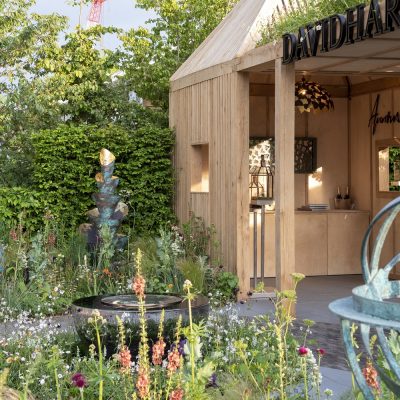 Wooden building in front of hedge with planted beds and round water feature on David Harber trade stand, Chelsea.