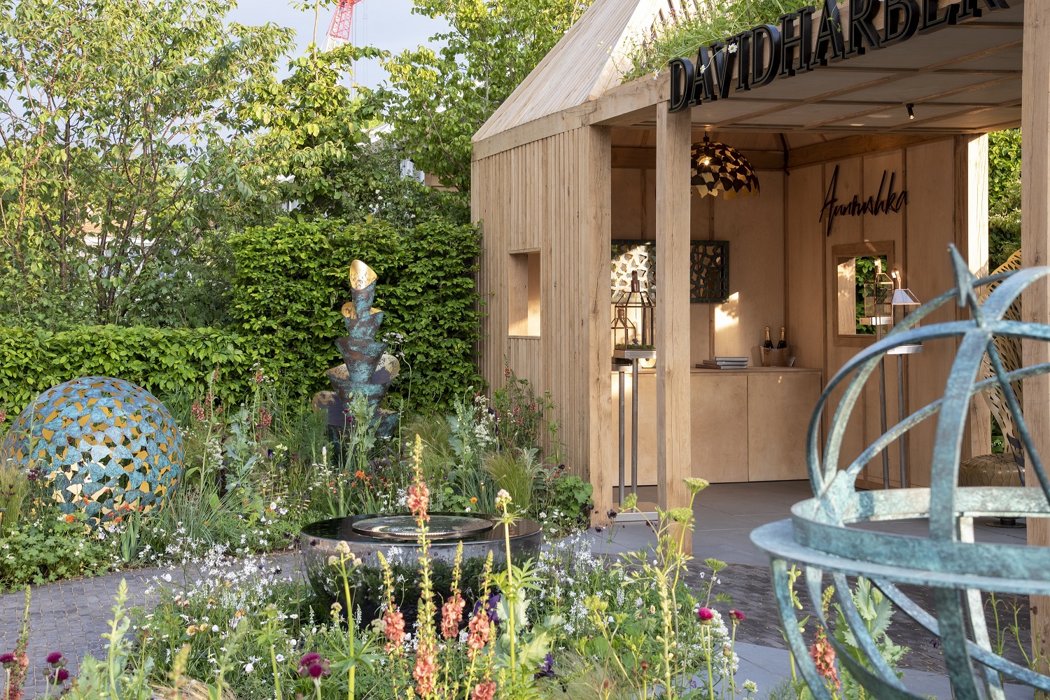 Wooden building in front of hedge with planted beds and round water feature on David Harber trade stand, Chelsea.