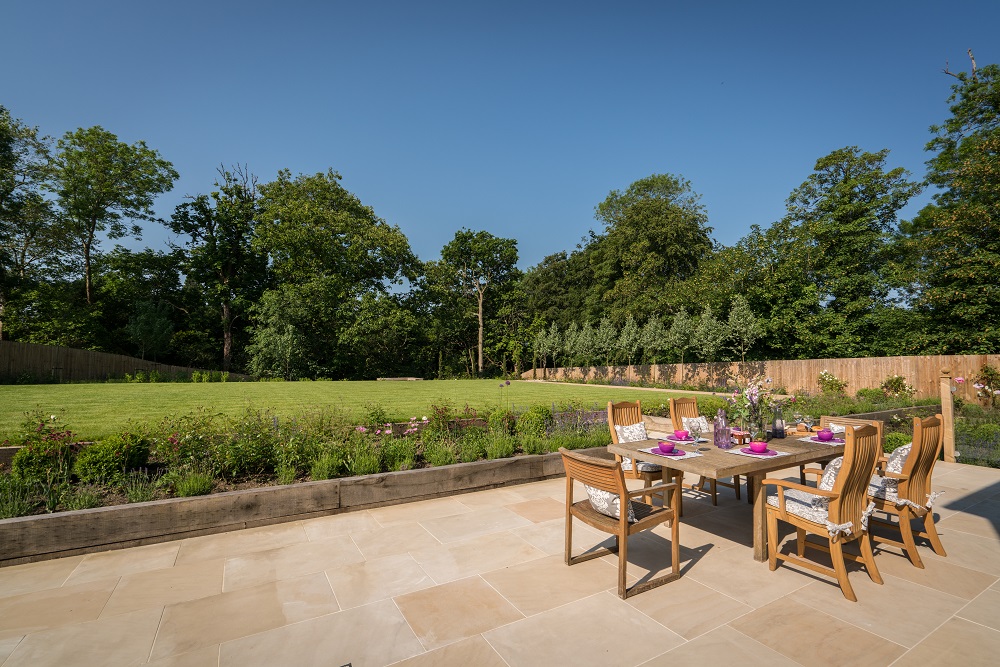 Harvest sawn sandstone patio with wooden outdoor furniture on edge of large lawn. Trees in background. 