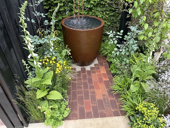 Tall pot water feature overflows onto gravel with apron of Dorset Antique clay pavers between relaxed planting
