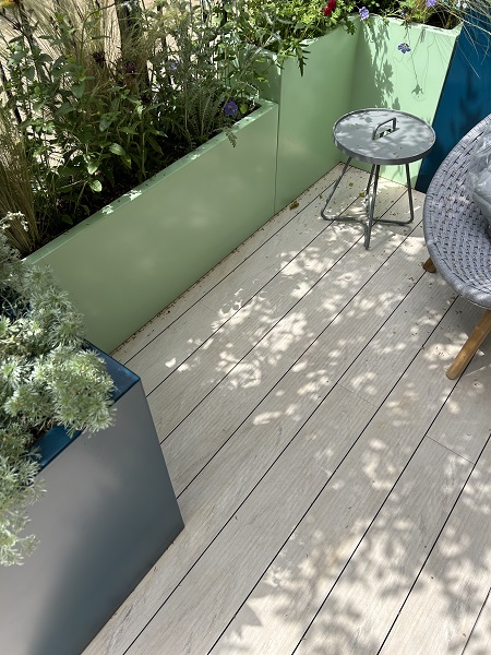 Dappled shade on DesignBoard Polar composite decking in RHS Chelsea 2023 balcony garden, with green and grey planters, small table and chair.