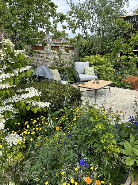 Outdoor chairs and table on Britannia Buff paving surrounded by verdant planting in The RSPCA Garden, Chelsea Flower Show 2023.