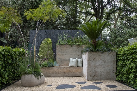 Large hypertufa containers against black slate cladding wall with tall mirror. Gravel and dark stepping stones in The Shifting Garden, RHS Chelsea 2023