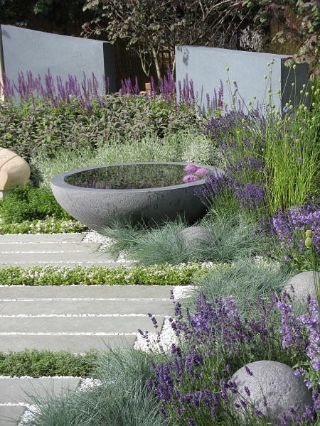 Large grey bowl water feature reflects purple blooms at far end of blue limestone  plank paving a gravel path.