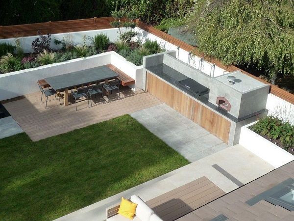 Outdoor living space with pizza oven, cupboards, raised beds and paving lining garden boundaries. Lawn and dining set in centre.