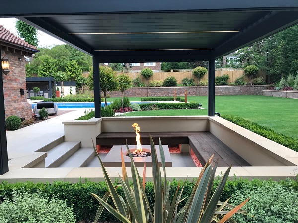 Lowered garden seating area with benches around fire pit under pergola. 
