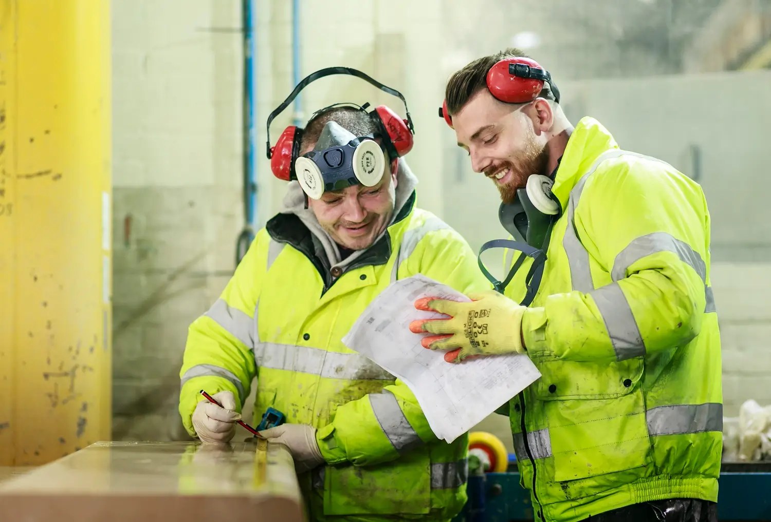 2 men in hi-vis jackets and ear defenders laugh as look at piece of paper one of them is holding.