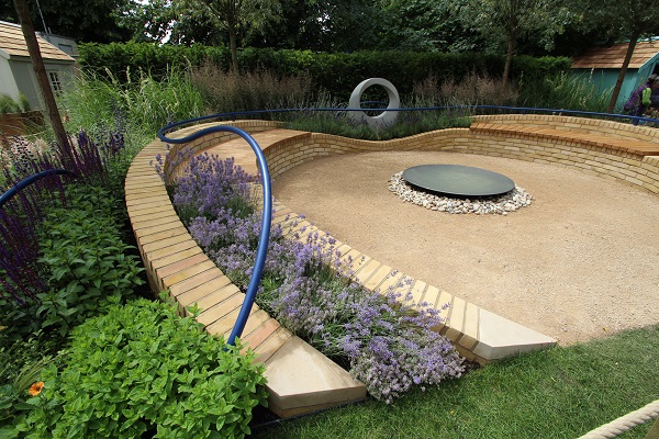 Curved raised bed leads to sculpture at apex of gravel area with shallow water bowl over pebbles. Hampton Court Flower Show 2016.