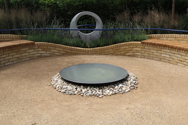Round shallow water bowl sits on smooth pebbles in middle of Breedon gravel area edged with curved brick raised bed. 