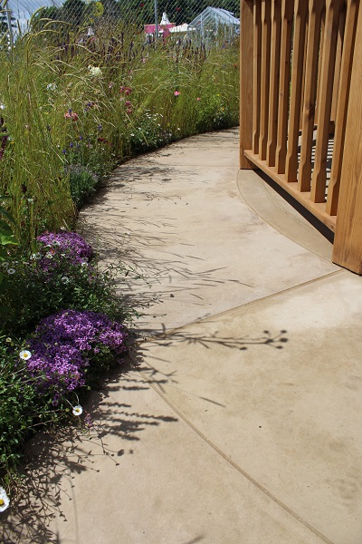 Path of Buff Yorkstone curves between wooden rails and bed planted with purple flowers.  Hampton Court Flower Show 2016. 