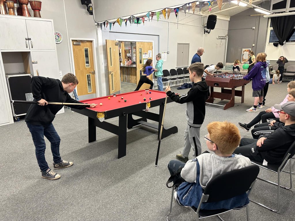Boys playing on small red-topped pool table in large room with others watching, in Crossroads Youth Club, Stone, Staffs.