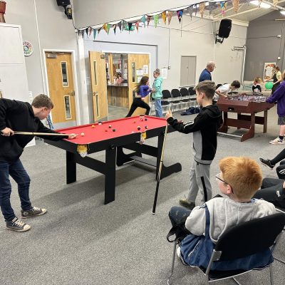 Boys playing on small red-topped pool table in large room with others watching, in Crossroads Youth Club, Stone, Staffs.