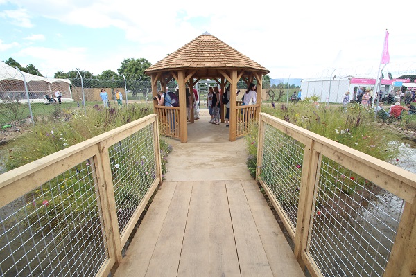 Wooden plank path leads between wire railings to Buff Yorkstone paving and octagonal roofed hut, UNHCR Border Garden 2016.