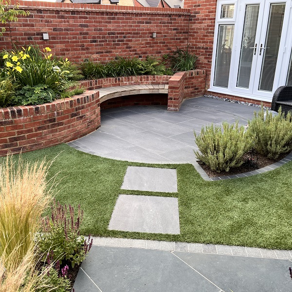 Curve-edged patio in walled garden. 2 slabs set into lawn lead to 2nd paved area. 