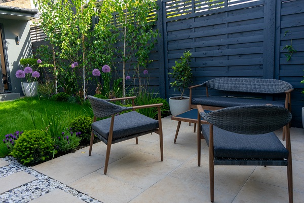 Planted border and lawn next to blue-cushioned garden sofa and chairs next to matching fence on Jura Beige porcelain patio.