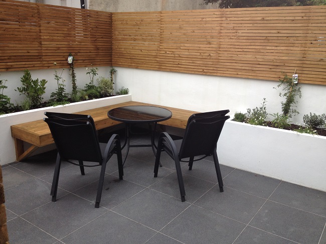 Walled and fenced corner of garden paved with Black Basalt vitrified porcelain slabs, with white raised beds and 2 chairs and table.