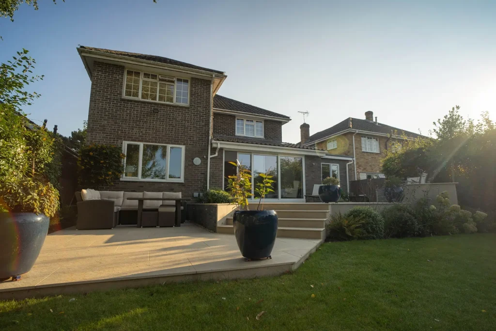 Patio outside glass extension of modern house descends to lower level via steps.