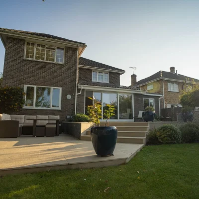 Patio outside glass extension of modern house descends to lower level via steps.