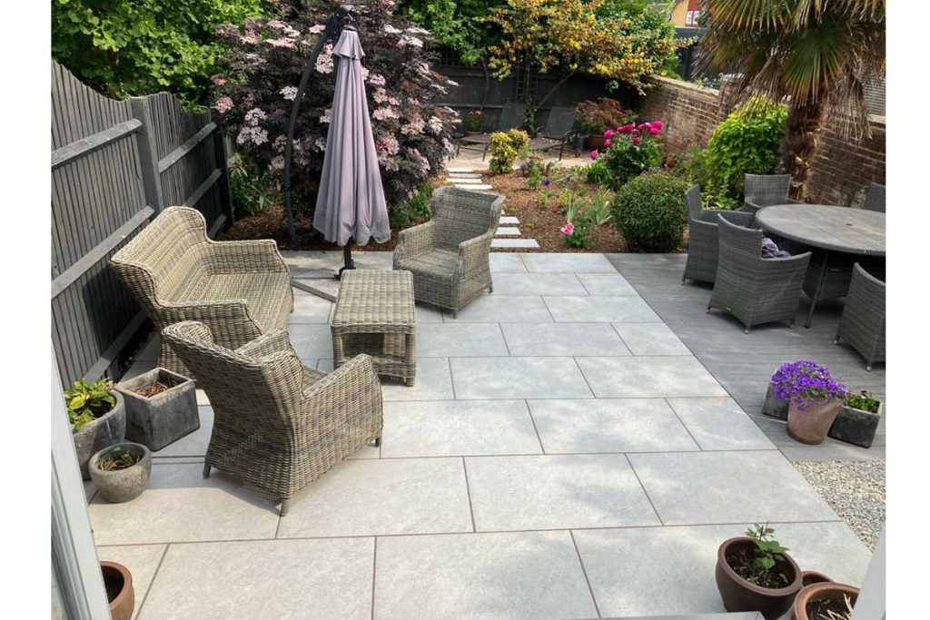 Long narrow rear garden with rattan furniture on Astor Grey porcelain patio, stepping stones through bed beyond.