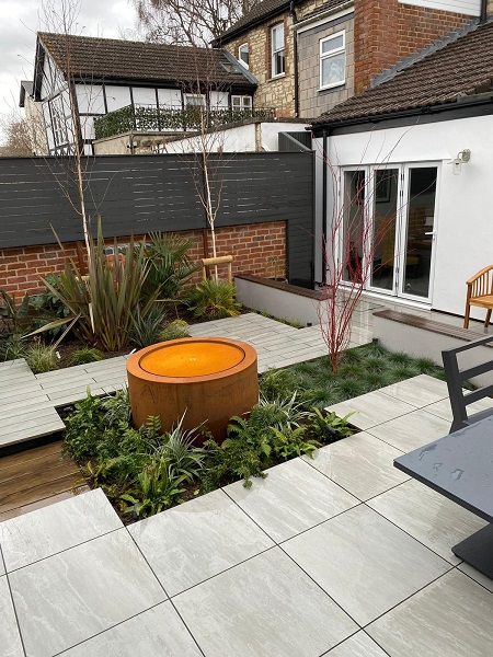 A patio of Kandla Grey 600x600mm porcelain slabs and plank paving with water feature in midst of planted bed.