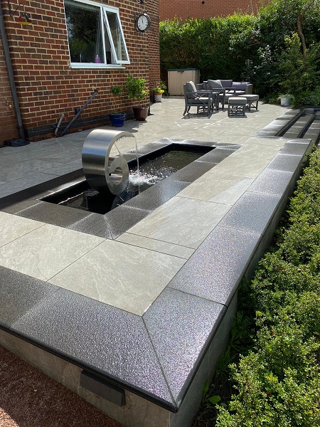 Kandla Grey porcelain patio with curled stainless steel water feature in narrow oblong pond edged with Black Granite.