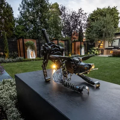 Metal caricature of dog on plinth faced with Steel Dark DesignClad in garden with large lawn and 3 large white planters against fence in background.
