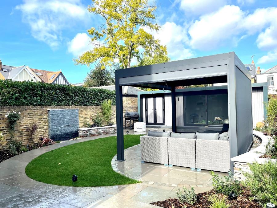 Pergola over an oval patio with path curving round lawn past wall water feature.