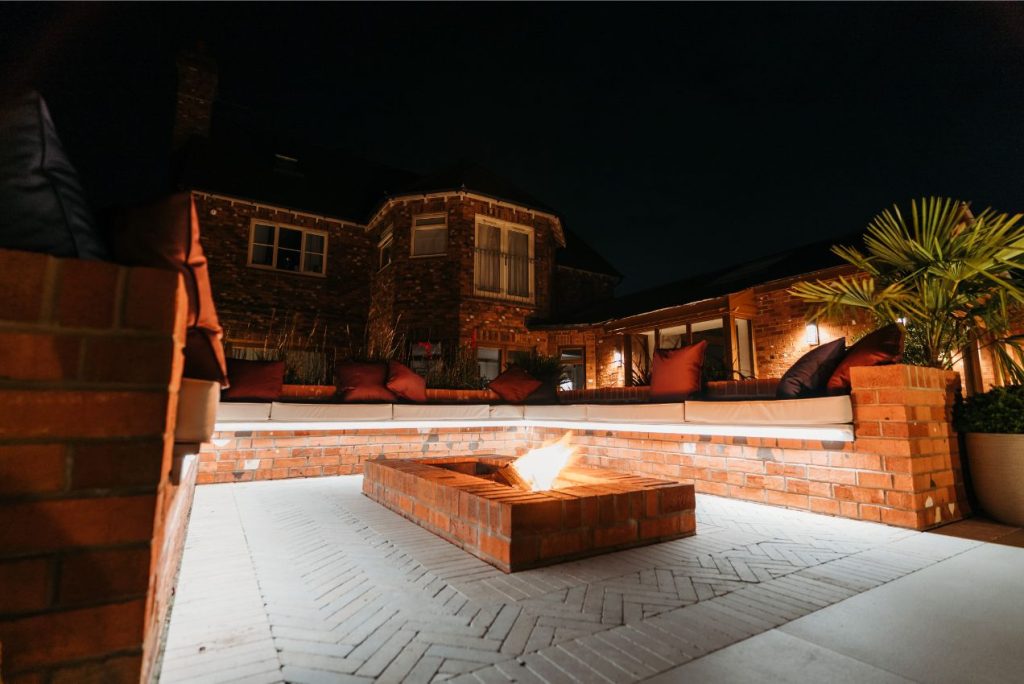 Illuminated night view of fire pit with in-built seating and Egyptian Beige stone paving. House behind.