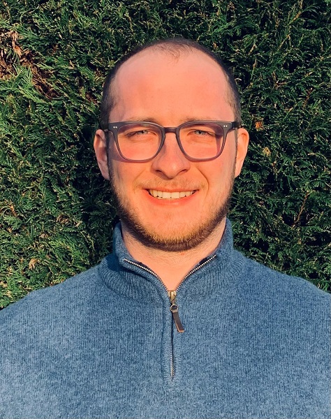 Man with glasses and partly bald head against evergreen background.