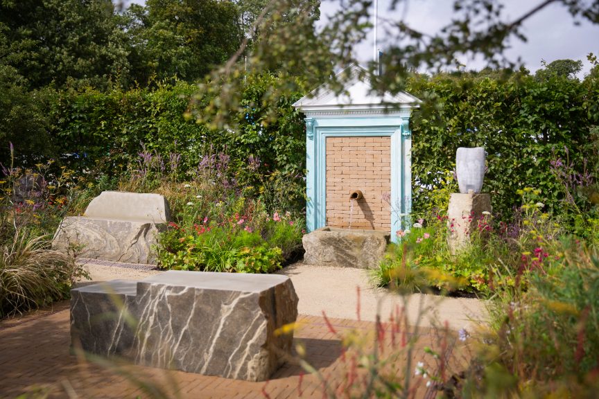 Water spout in brick wall in door frame with Golden Buff clay pavers, naturalistic planting and rock seating.