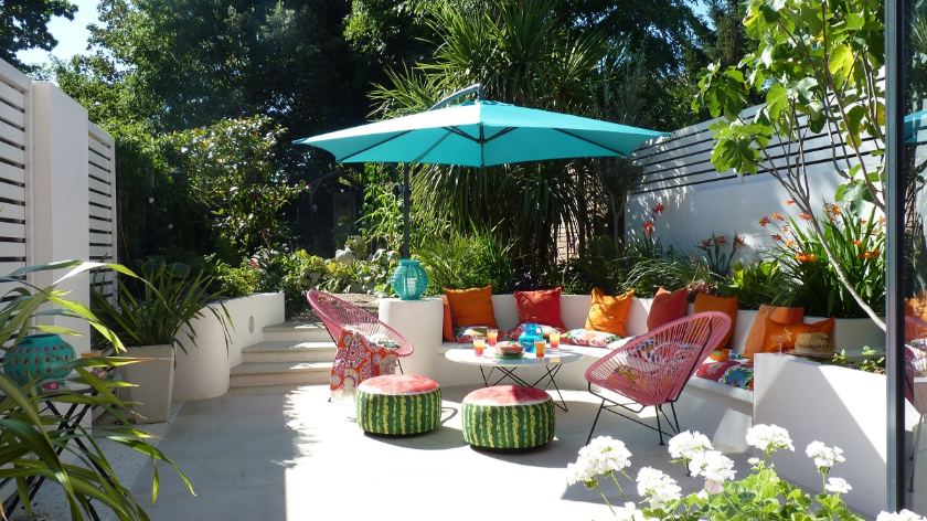 Back garden patio in Beige sawn sandstone, wtih built in bench and colourful cushions at parasol covered table.